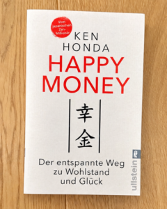 Das Buch „Happy Money“ von Ken Honda auf einem Holzboden